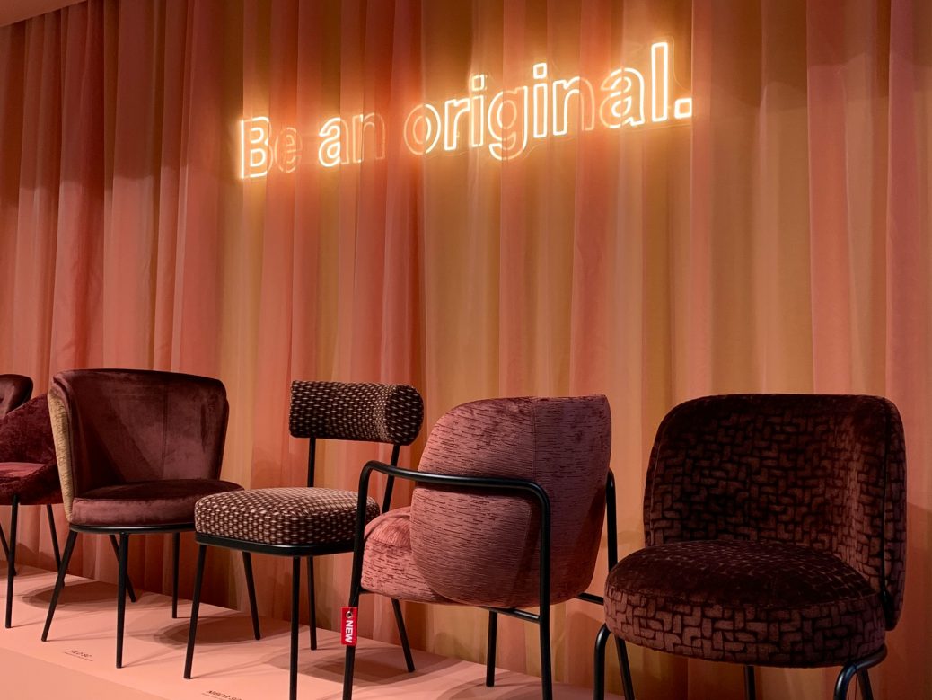 Ausstellung moderner Designerstühle in warmen Beerentönen vor einem Vorhang mit Leuchtschrift ‚Be an original‘ – kreative Möbelszene auf der Salone del Mobile 2025.